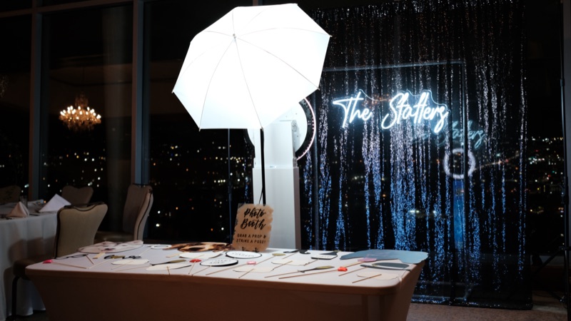 An open-air photo booth with a lit umbrella, props table and sequin backdrop at a reception.