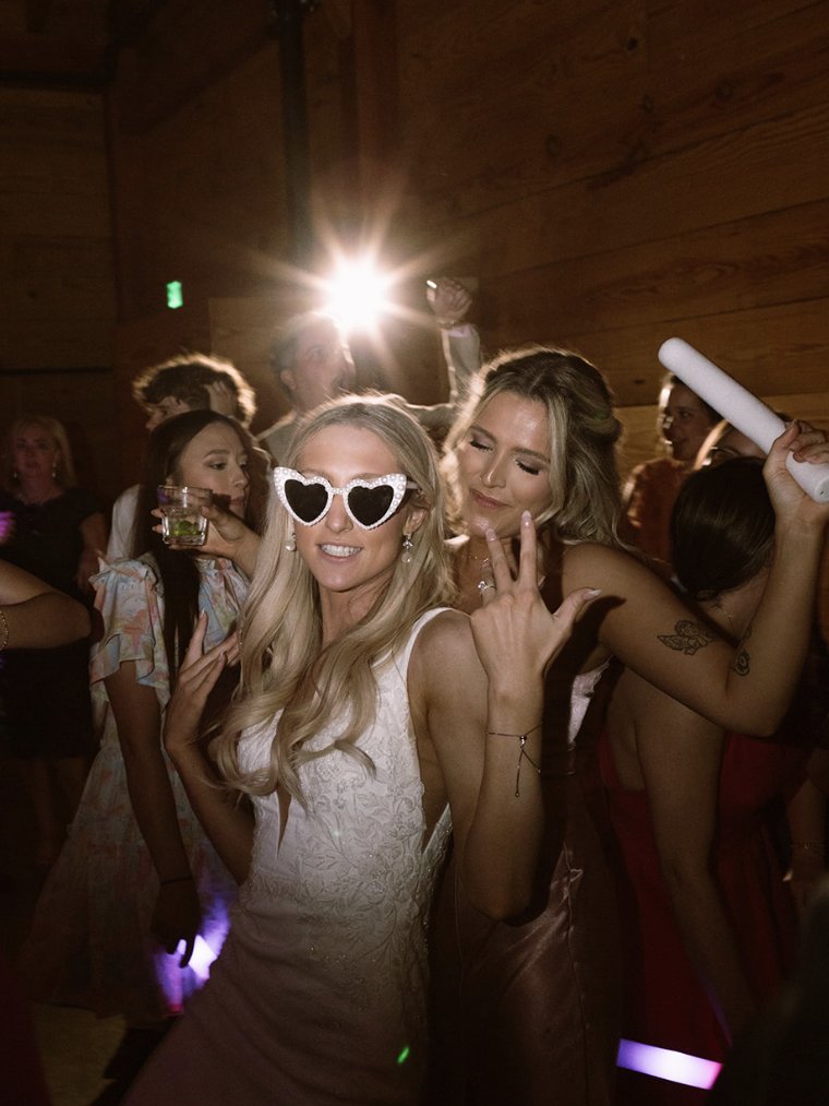 A bride in heart-shaped sunglasses dancing with a friend on a crowded dance floor.